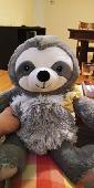 A grey sloth toy with a fluffy body and a yellow velvet scrunchie on the arm.